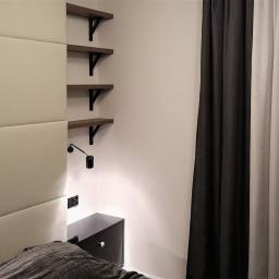 MAGURA - Sypialnia z tapicerowanym panelem ściennym, trzema drewnianymi półkami na czarnych wspornikach, czarną szafką nocną, lampką i zasłonami w odcieniach szarości.