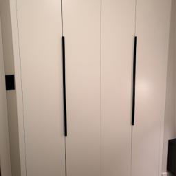 MAGURA - Biała szafa wnękowa z czterema drzwiami i długimi, pionowymi, czarnymi uchwytami, widoczna z bliska, z fragmentem podłogi i szafki nocnej w tle.