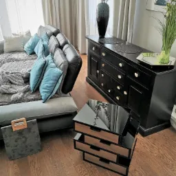 Salon z szarym narożnikiem, turkusowymi poduszkami, czarną komodą z jasnymi gałkami i drewnianym stolikiem kawowym z lustrzanym blatem.