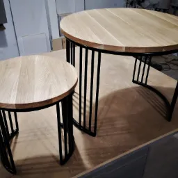 Dwa okrągłe stoliki kawowe z blatem z jasnego drewna i czarnymi, metalowymi nogami w kształcie pionowych prętów, ustawione na blacie roboczym w warsztacie stolarskim.