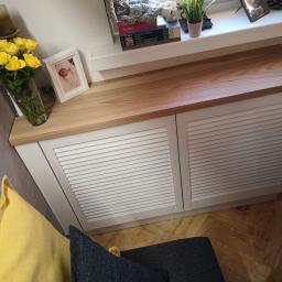 MAGURA - Biała szafka z lamelowymi drzwiami i blatem z jasnego drewna, pełniąca funkcję zabudowy grzejnika pod oknem, z dekoracjami na blacie: wazon z żółtymi różami, ramka ze zdjęciem, książki o sztuce...