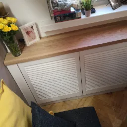 Biała szafka z lamelowymi drzwiami i blatem z jasnego drewna, pełniąca funkcję zabudowy grzejnika pod oknem, z dekoracjami na blacie: wazon z żółtymi różami, ramka ze zdjęciem, książki o sztuce...