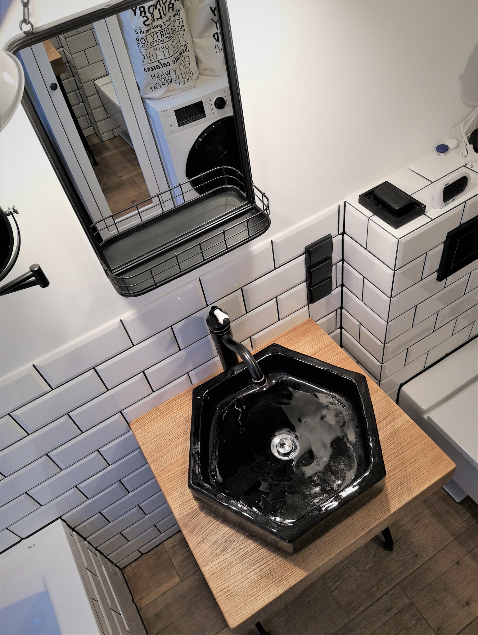 Czarna, sześciokątna umywalka nablatowa z czarną baterią, ustawiona na blacie z jasnego drewna, w nowoczesnej łazience z białymi płytkami imitującymi cegłę. W lustrze odbija się pralka i torba...