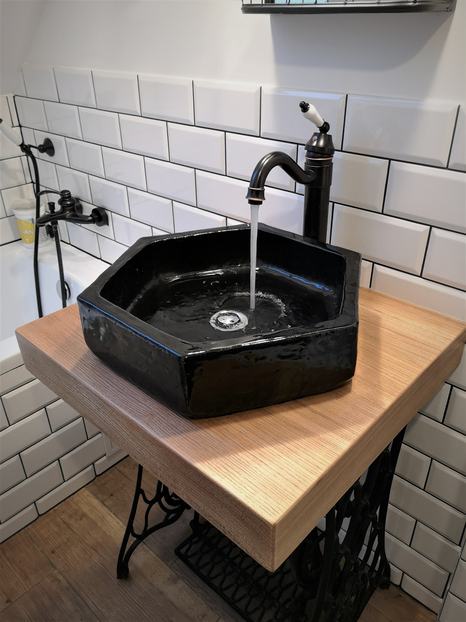 Czarna, ośmiokątna umywalka nablatowa z chromowanym odpływem i czarną baterią, zamontowana na drewnianym blacie wspartym na metalowej konstrukcji maszyny do szycia, w łazience z białymi płytkami...