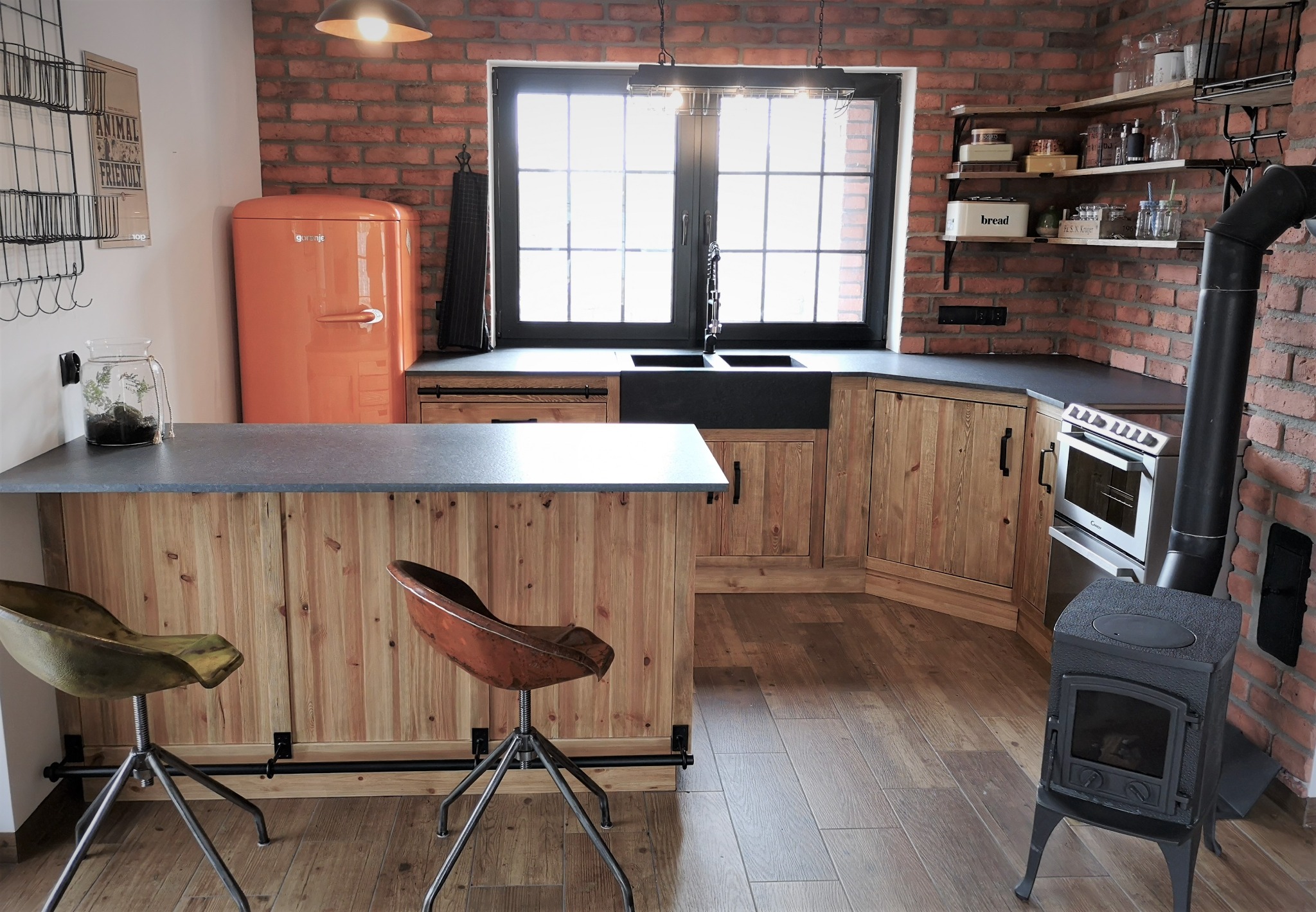 Rustykalna kuchnia z ceglaną ścianą, drewnianymi szafkami, czarnym blatem, pomarańczową lodówką retro i piecem wolnostojącym.