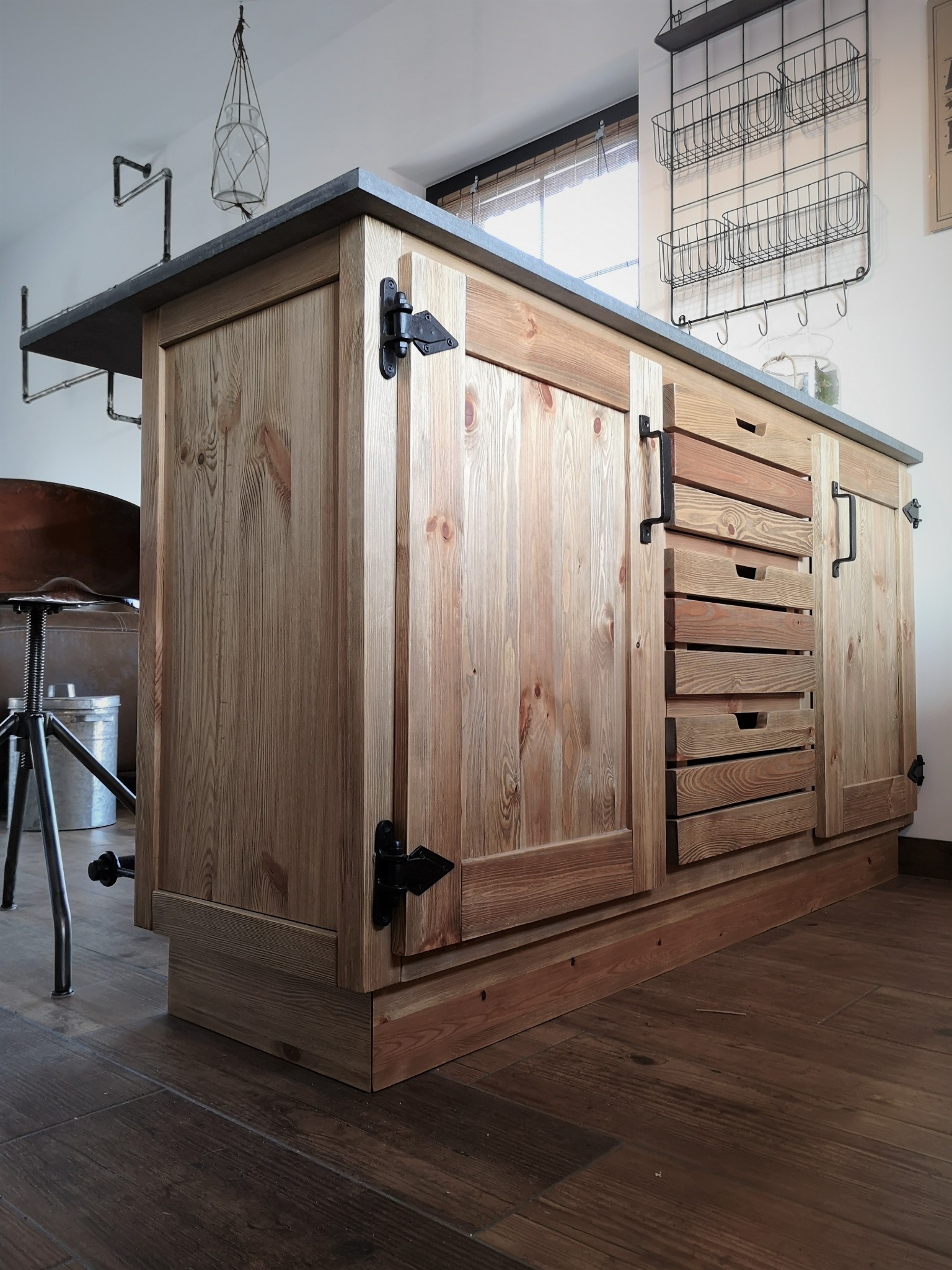 Rustykalna wyspa kuchenna z surowego drewna z czarnymi okuciami i szarym blatem, widoczna na tle drewnianej podłogi i metalowego stołka barowego.