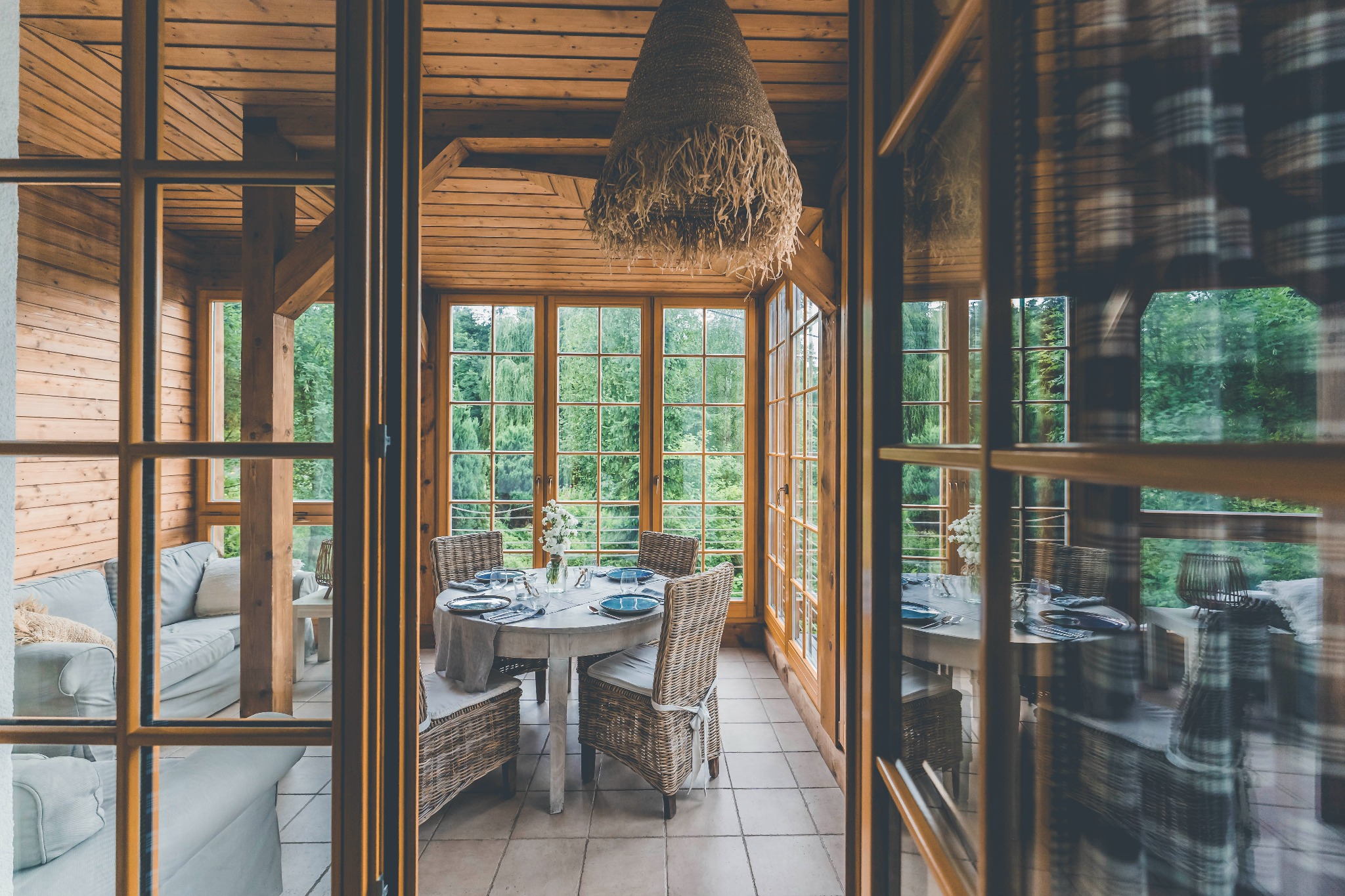 Jasna oranżeria z drewnianą konstrukcją, okrągłym stołem nakrytym dla czterech osób z niebieską zastawą i wiklinowymi krzesłami, widok przez przeszklone drzwi.