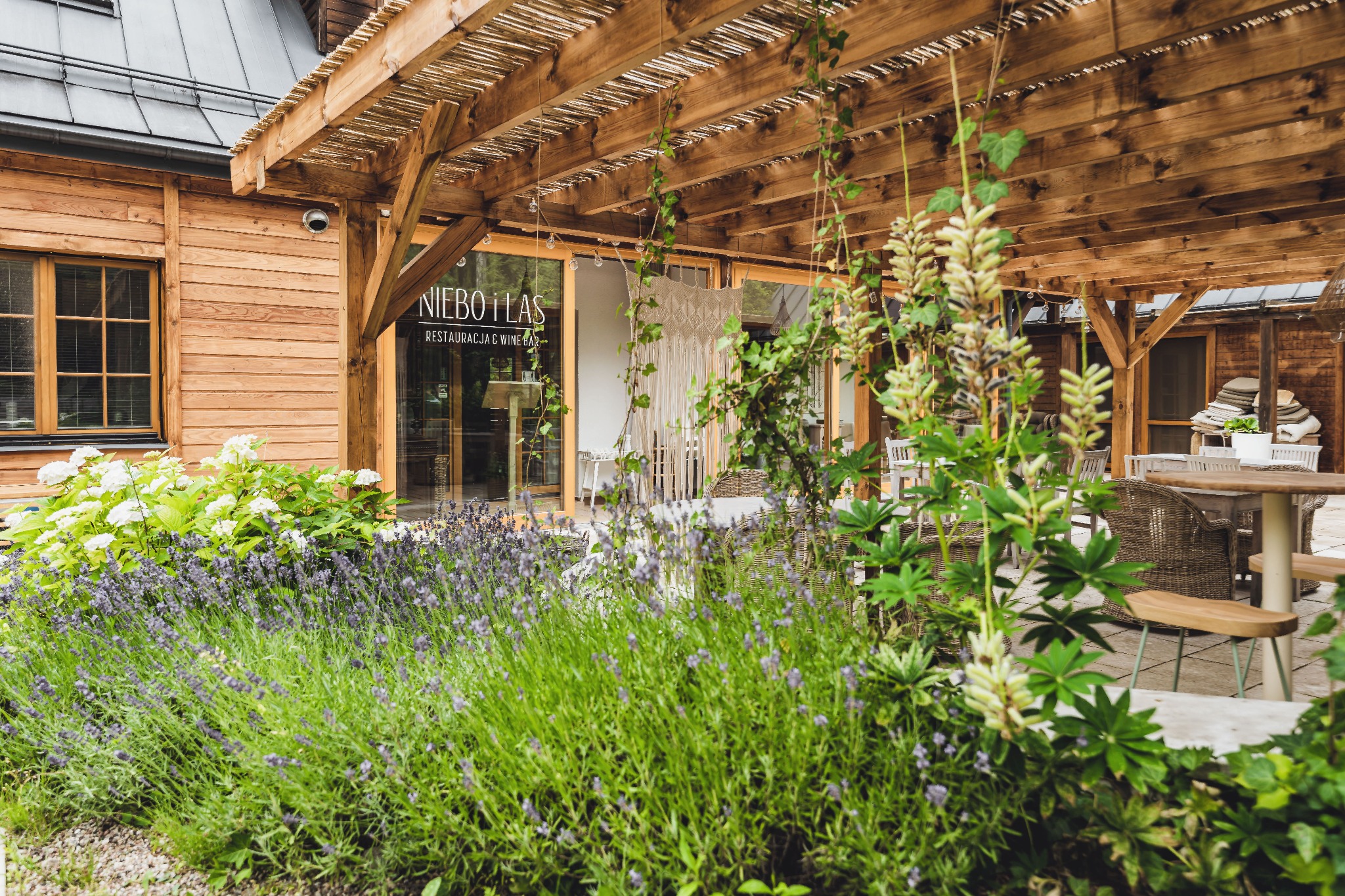 Zewnętrzny widok restauracji z drewnianą elewacją, patio z pergolą porośniętą roślinami, stolikami i krzesłami z wikliny oraz rabatą lawendy i hortensji.
