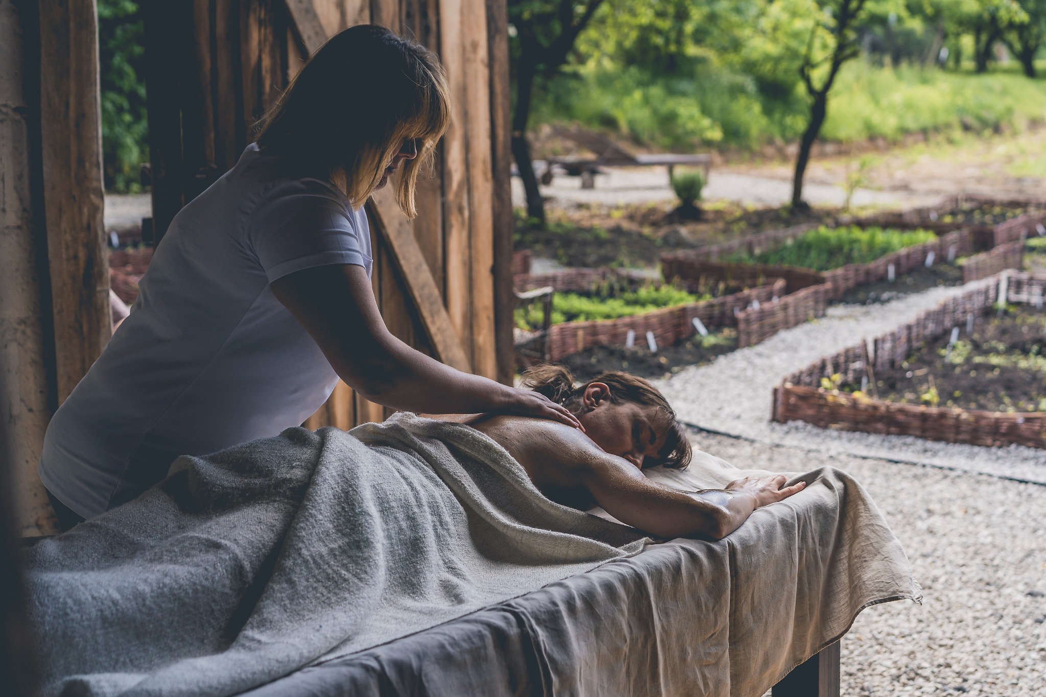 Kobieta otrzymuje masaż pleców w rustykalnym otoczeniu, widoczny ogród z drewnianymi płotkami w tle.