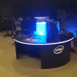 Okrągłe, czarne biurko gamingowe z podświetleniem LED, komputerem NTT i logo Intel, ustawione na tle akustycznej ściany w ciemnym pomieszczeniu.