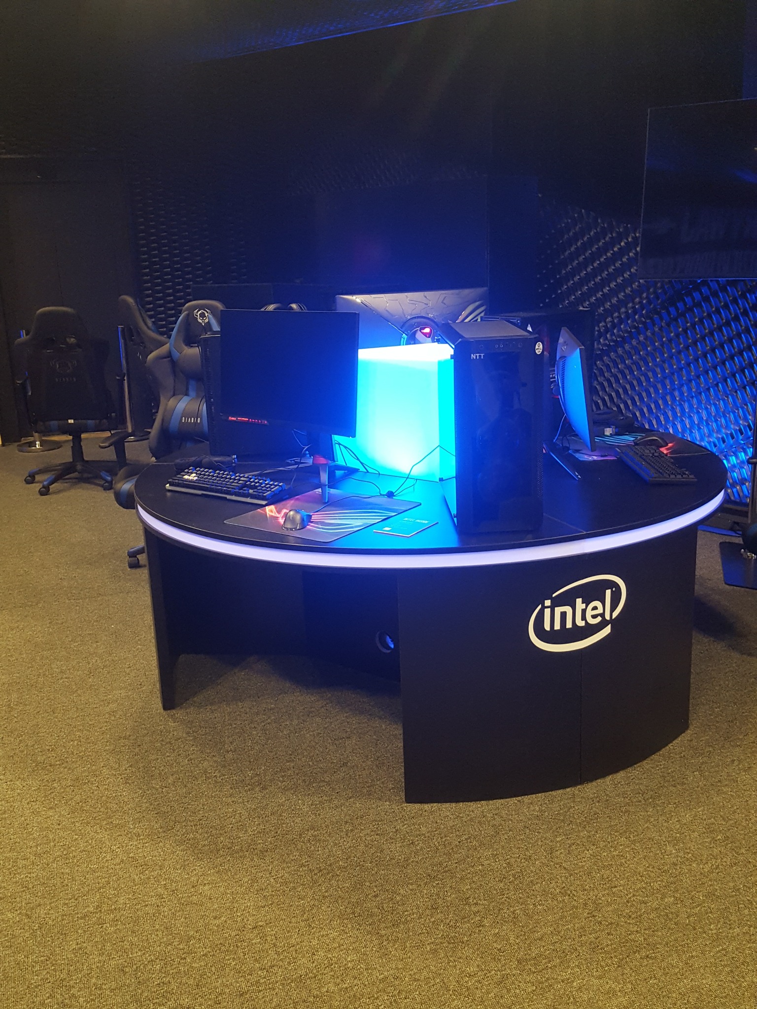 Okrągłe, czarne biurko gamingowe z podświetleniem LED, komputerem NTT i logo Intel, ustawione na tle akustycznej ściany w ciemnym pomieszczeniu.