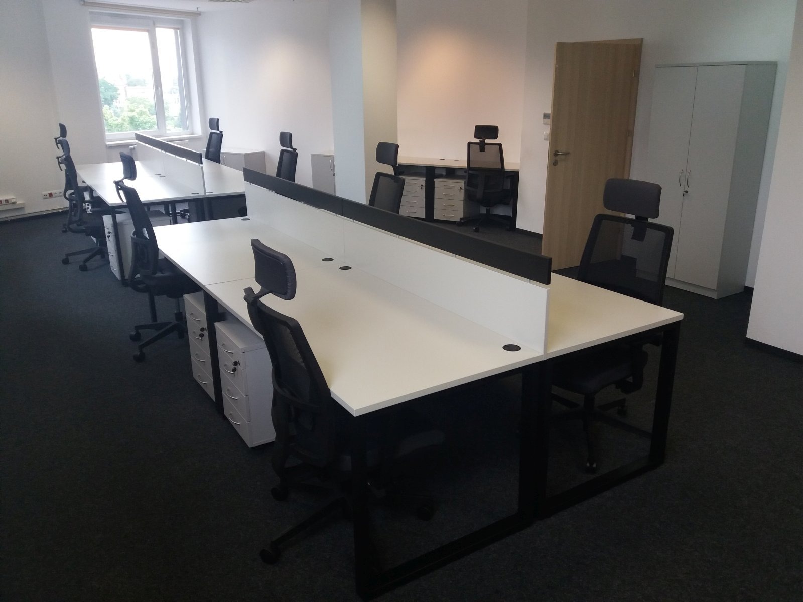 Nowoczesne biuro z białymi biurkami z czarnymi nogami i przegrodami, czarne krzesła biurowe, białe szafki na dokumenty, jasne wnętrze z dużym oknem.