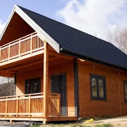 Nowoczesny dom szkieletowy z drewnianą elewacją, czarnym dachem dwuspadowym i balkonem z drewnianą balustradą, widoczny na tle drzew i błękitnego nieba z chmurami.