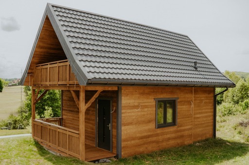 Gotowy, dwukondygnacyjny budynek z drewna z balkonem, pokryty szarą blachodachówką, na tle zieleni.