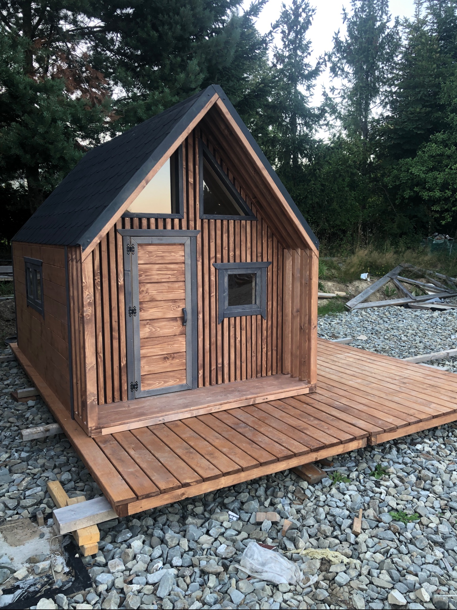 Mały, nowoczesny domek z drewna z czarnym dachem i drewnianym tarasem, osadzony na kamienistym podłożu, otoczony zielenią.