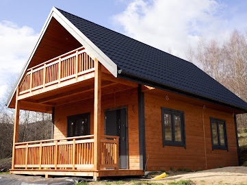 Nowoczesny dom szkieletowy z drewnianą elewacją, czarnym dachem dwuspadowym i balkonem z drewnianą balustradą, widoczny na tle drzew i błękitnego nieba z chmurami.