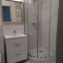 Nowoczesna łazienka z kabiną prysznicową narożną, białą szafką pod umywalką i lustrem w drewnianej ramie, wykończona jasnymi płytkami.