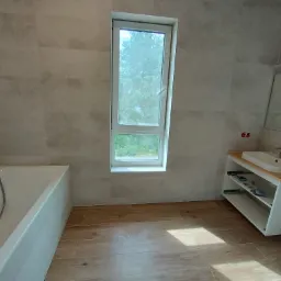 Jasna łazienka z beżowymi płytkami imitującymi kamień, drewnopodobna podłoga, biała wanna, szafka z umywalką i wąskie okno z widokiem na zieleń.