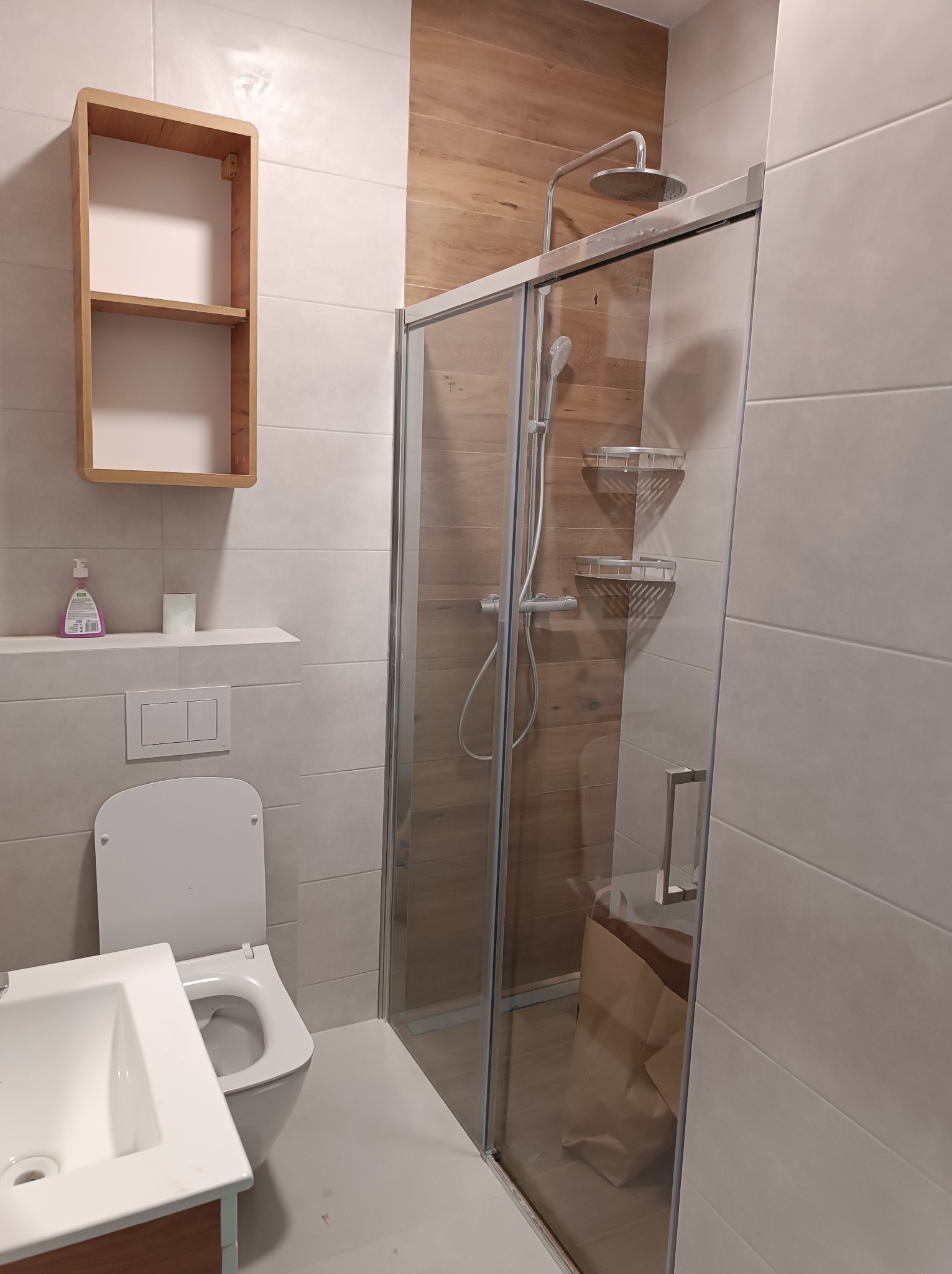 Nowoczesna łazienka z kabiną prysznicową typu walk-in, drewnianymi akcentami na ścianie i otwartą półką z jasnego drewna, biała ceramika sanitarna i płytki w odcieniach szarości.