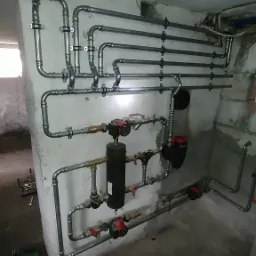 Złożony system rur grzewczych z pompami obiegowymi i zaworami, zamontowany na ścianie w piwnicy z widocznymi narzędziami instalacyjnymi na podłodze.