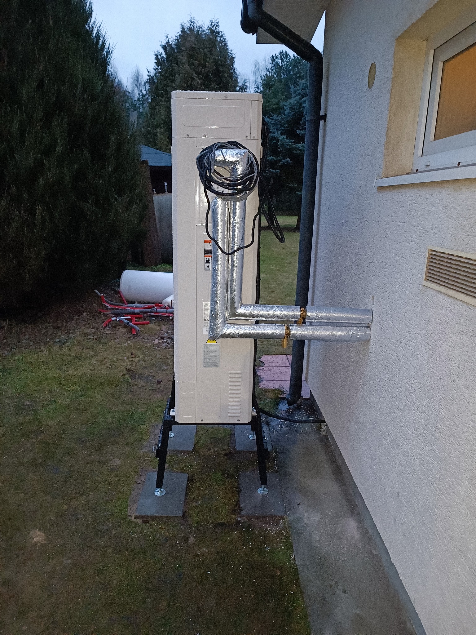 Biała pompa ciepła typu monoblok, zainstalowana na zewnątrz budynku na czarnych wspornikach, z podłączonymi izolowanymi rurami w srebrnej folii, widoczne przewody elektryczne, w tle trawnik, drzewa...
