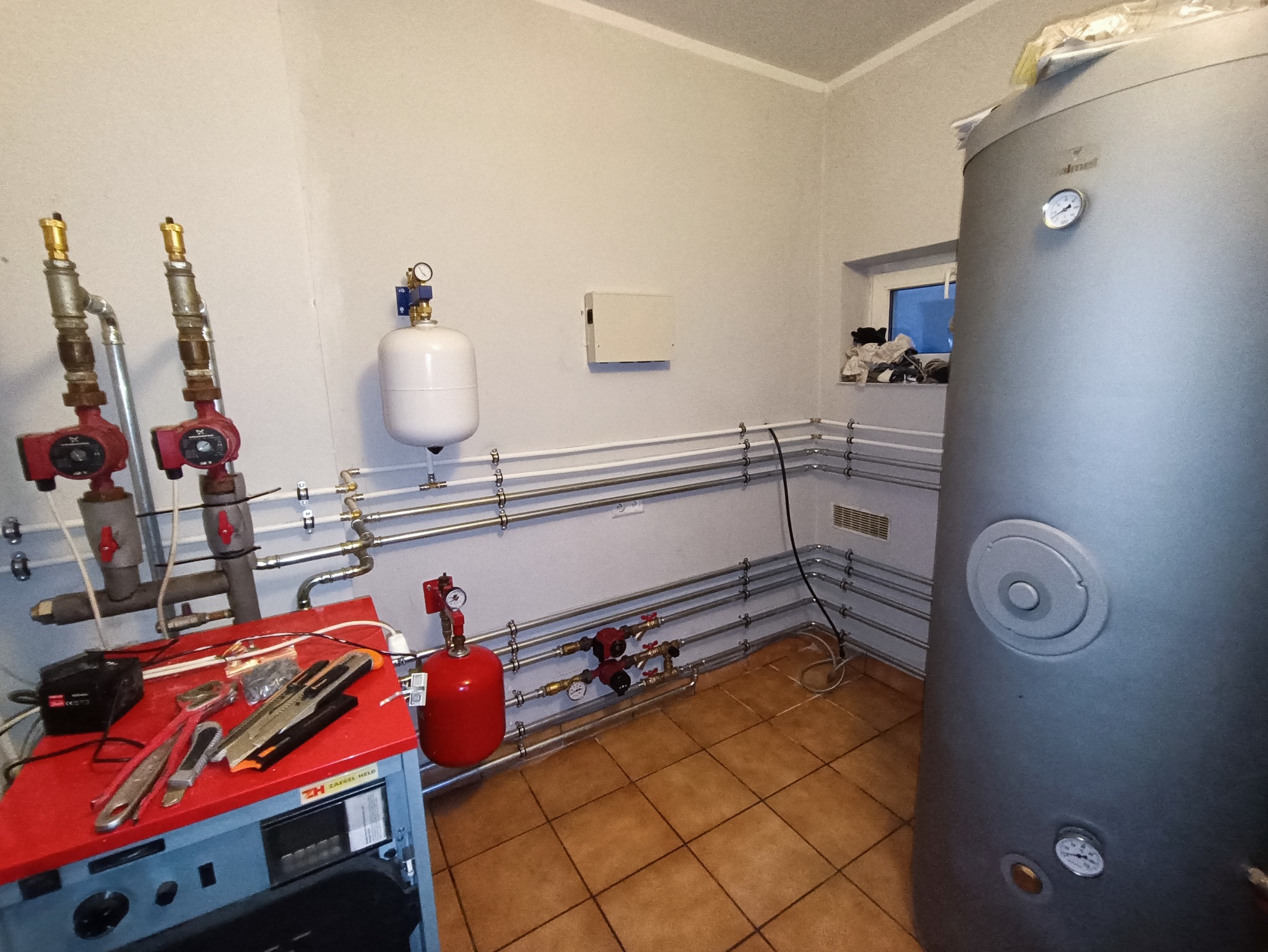Wnętrze kotłowni z szarym zbiornikiem akumulacyjnym, czerwonymi pompami obiegowymi, białym naczyniem wzbiorczym, licznymi rurami, narzędziami na czerwonym blacie i brązową podłogą z płytek.