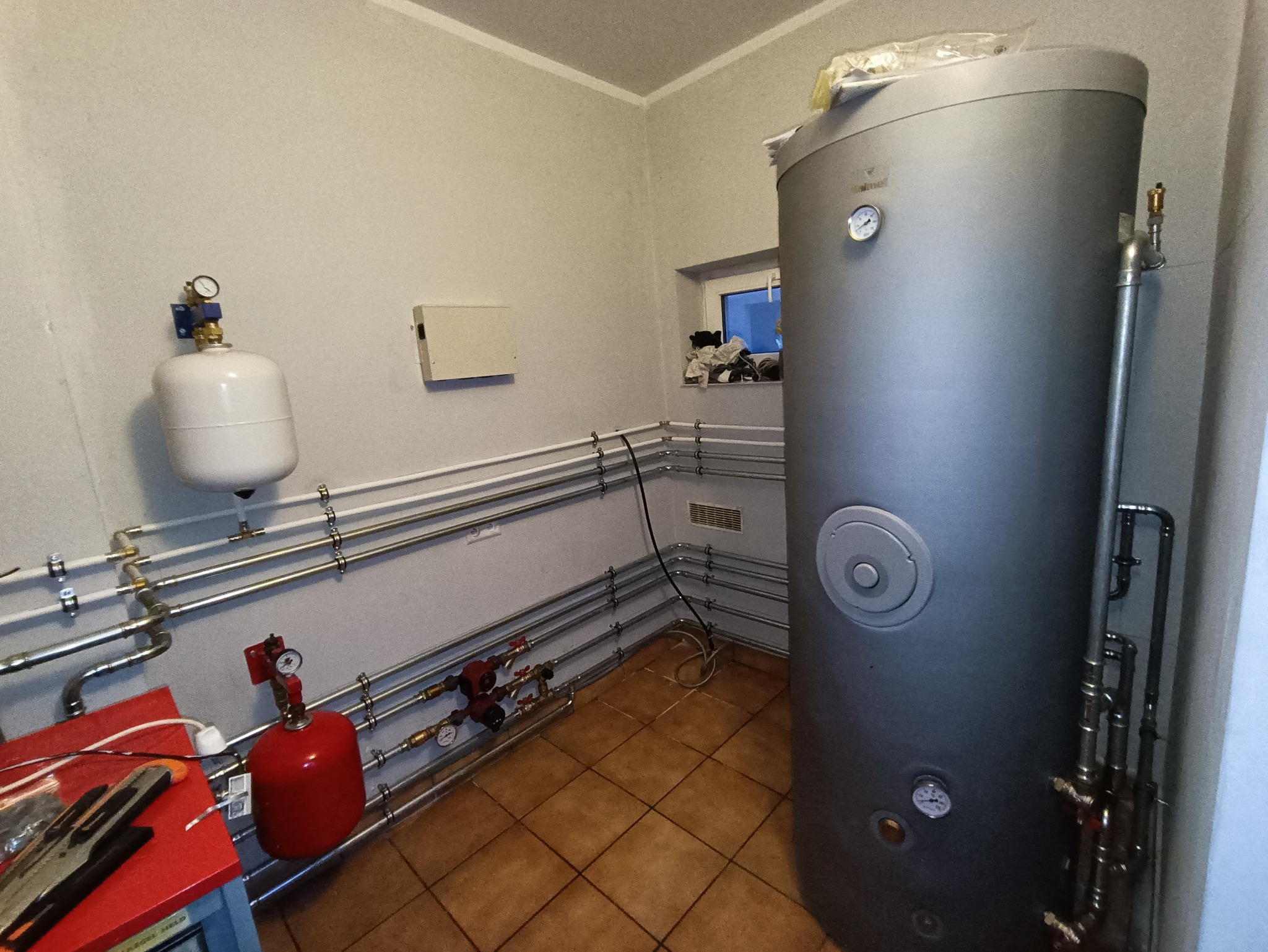 W kotłowni, szary, cylindryczny zbiornik akumulacyjny ciepła z manometrami, podłączony do systemu rur miedzianych i białych, z widocznymi zaworami, naczyniem przeponowym i narzędziami na czerwonym...