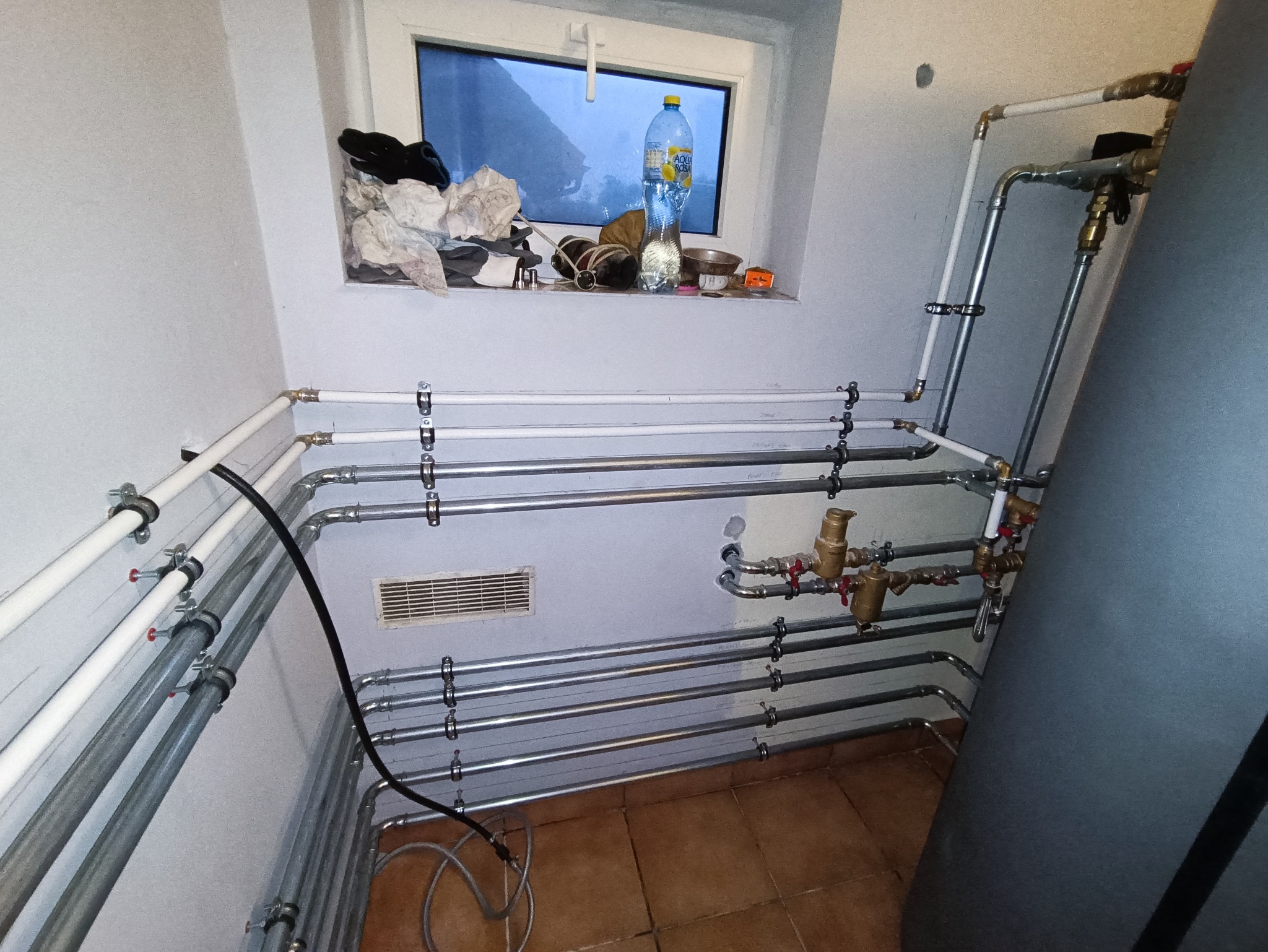 Kompleksowa instalacja hydrauliczna z licznymi rurami miedzianymi i białymi, zaworami oraz czarnym przewodem elektrycznym, umieszczona w pomieszczeniu z brązowymi płytkami podłogowymi i oknem...