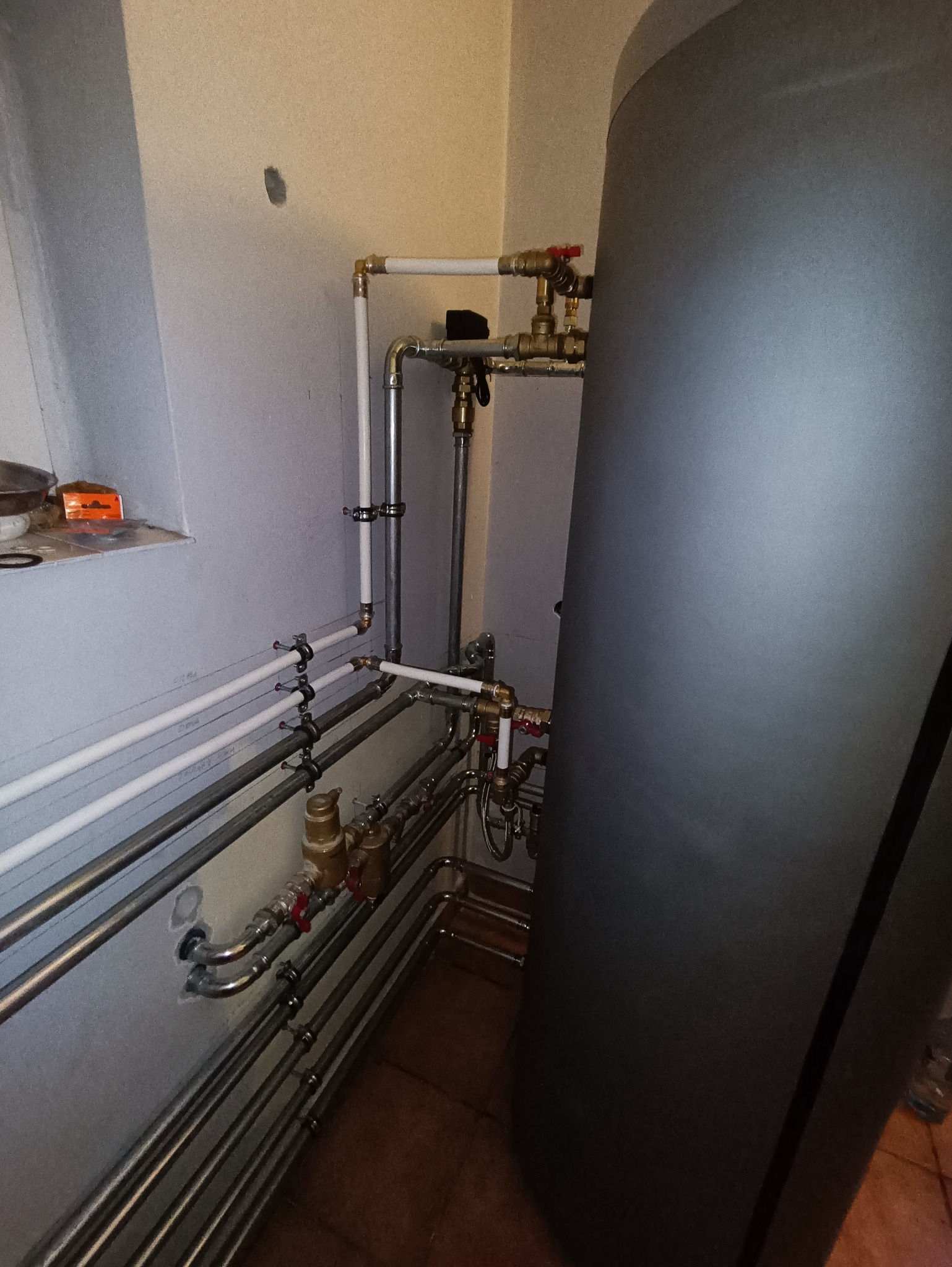 Szczegółowe ujęcie instalacji hydraulicznej z rurami miedzianymi i plastikowymi, zaworami z czerwonymi pokrętłami oraz szarym zbiornikiem akumulacyjnym w ciasnym pomieszczeniu technicznym, widoczne...