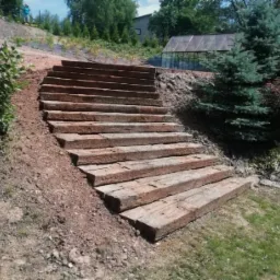 Schody z drewnianych belek na zboczu, prowadzące w górę ogrodu z widoczną szklarnią w tle.