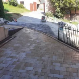 Ułożona kostka brukowa w odcieniach beżu i brązu z dekoracyjnym obramowaniem, prowadząca do bramki z kutego żelaza i dalej do podjazdu z ciemniejszej kostki; widoczna zagęszczarka w tle.