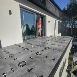 Układanie szarych płytek na tarasie domu jednorodzinnego z użyciem systemu poziomowania płytek, widoczny glazurnik w drzwiach balkonowych.