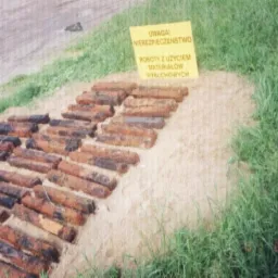 Odkryte, skorodowane pociski artyleryjskie ułożone rzędami na piaszczystym terenie, z ż&oacute;łtą tablicą ostrzegawczą o niebezpieczeństwie użycia materiał&oacute;w wybuchowych w tle.