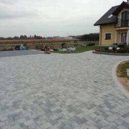 ENKM-BUD - Usługi remontowo budowlane - Rozległy podjazd z szarej kostki brukowej ułożonej we wzór jodełki, prowadzący do domu z żółtą elewacją i balkonem; w tle widoczne prace ziemne i krajobraz polny.