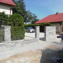 ENKM-BUD - Usługi remontowo budowlane - Budowa ogrodzenia z szarej kostki betonowej, pracownik wyrównuje podłoże pod bramą wjazdową, w tle domy z czerwonymi dachami i srebrny samochód.