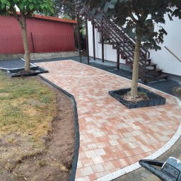 ENKM-BUD - Usługi remontowo budowlane - Kaskadowa ścieżka z kostki brukowej w odcieniach czerwieni i beżu, obramowana czarnym obrzeżem, wijąca się między drzewami i prowadząca do schodów zewnętrznych.