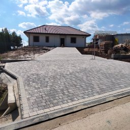 ENKM-BUD - Usługi remontowo budowlane - Szeroki podjazd z szarej kostki brukowej prowadzący do domu z czerwoną dachówką. Widoczne obrzeża i krawężniki. W tle budowa drugiego budynku.