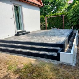 ENKM-BUD - Usługi remontowo budowlane - Nowoczesny taras z kostki brukowej w odcieniach szarości i czerni, z trzema stopniami i betonową obudową rabaty, przylegający do białego budynku z oknem.