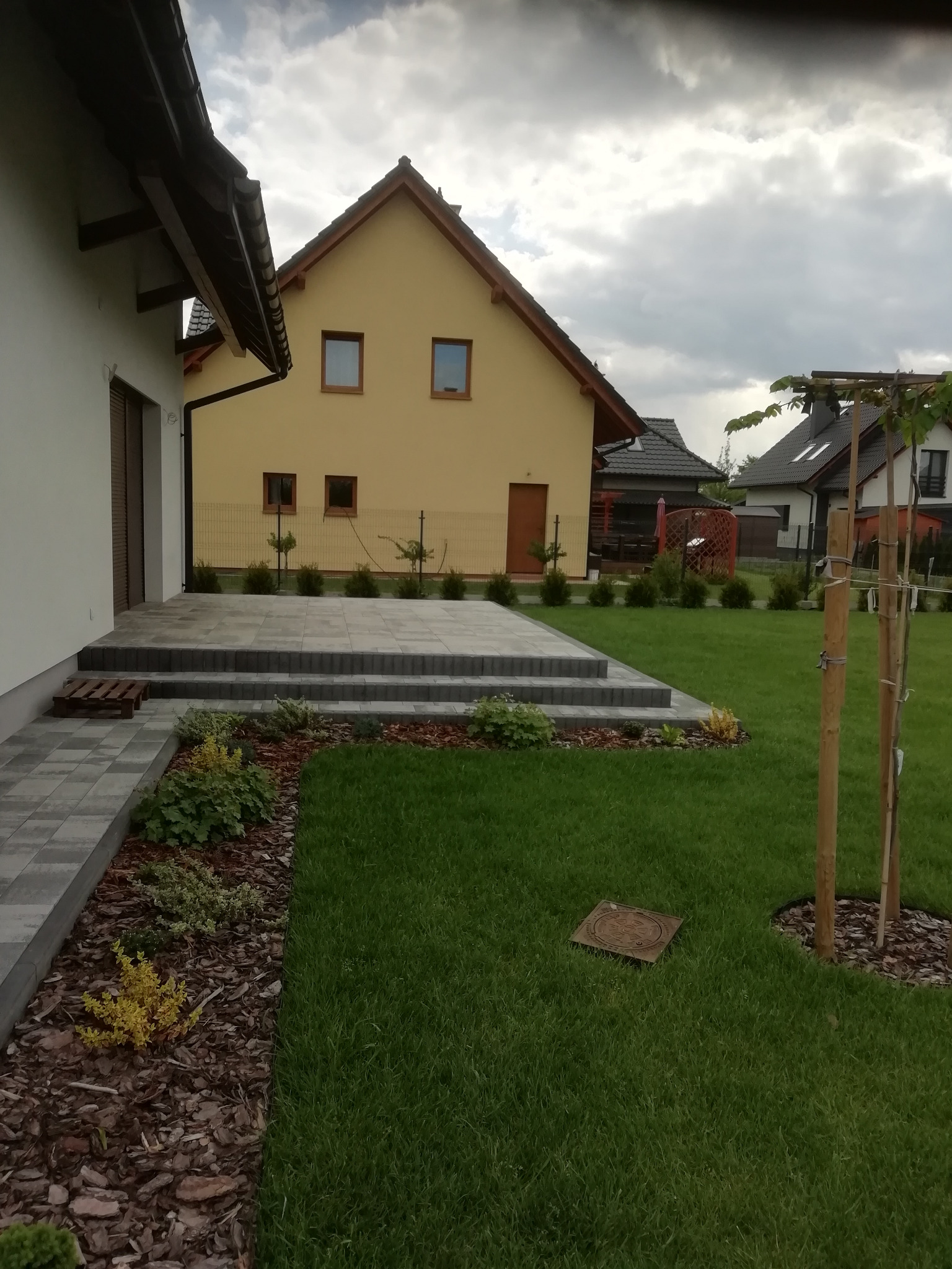 Nowoczesny taras z szarej kostki brukowej z trzema stopniami, obrzeża rabat wysypane korą, trawnik, w tle żółty dom i fragmenty innych budynków.