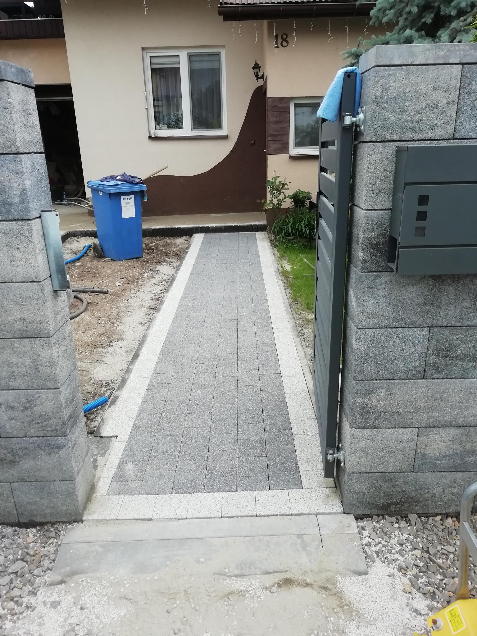 Ułożona kostka brukowa tworząca ścieżkę do bramy z szarej kostki, widoczny fragment domu z numerem 18 i niebieski pojemnik na śmieci.