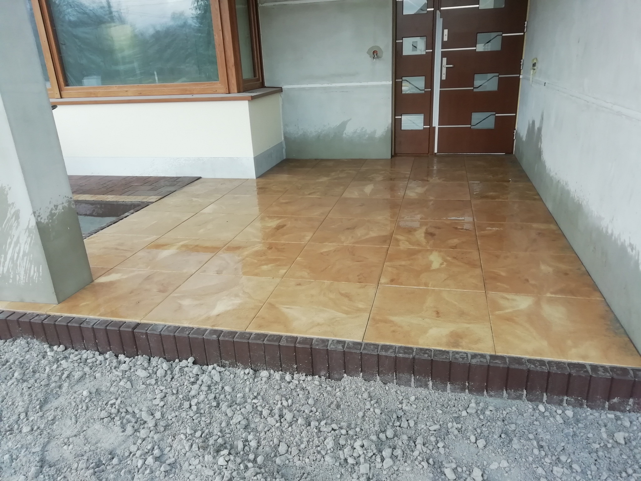 Wyłożony płytkami taras przed wejściem do domu, obrzeże z ciemnej kostki, podsypka z kruszywa, ściany domu w trakcie wykończenia.