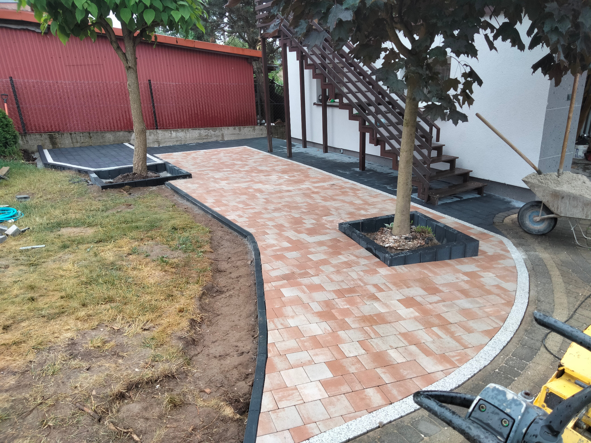 Kaskadowa ścieżka z kostki brukowej w odcieniach czerwieni i beżu, obramowana czarnym obrzeżem, wijąca się między drzewami i prowadząca do schodów zewnętrznych.