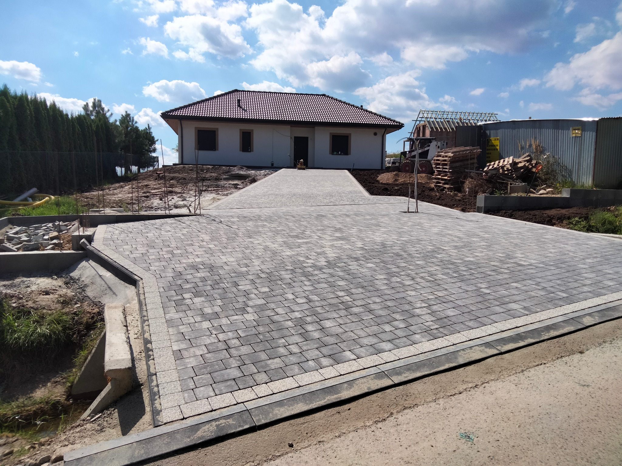 Szeroki podjazd z szarej kostki brukowej prowadzący do domu z czerwoną dachówką. Widoczne obrzeża i krawężniki. W tle budowa drugiego budynku.