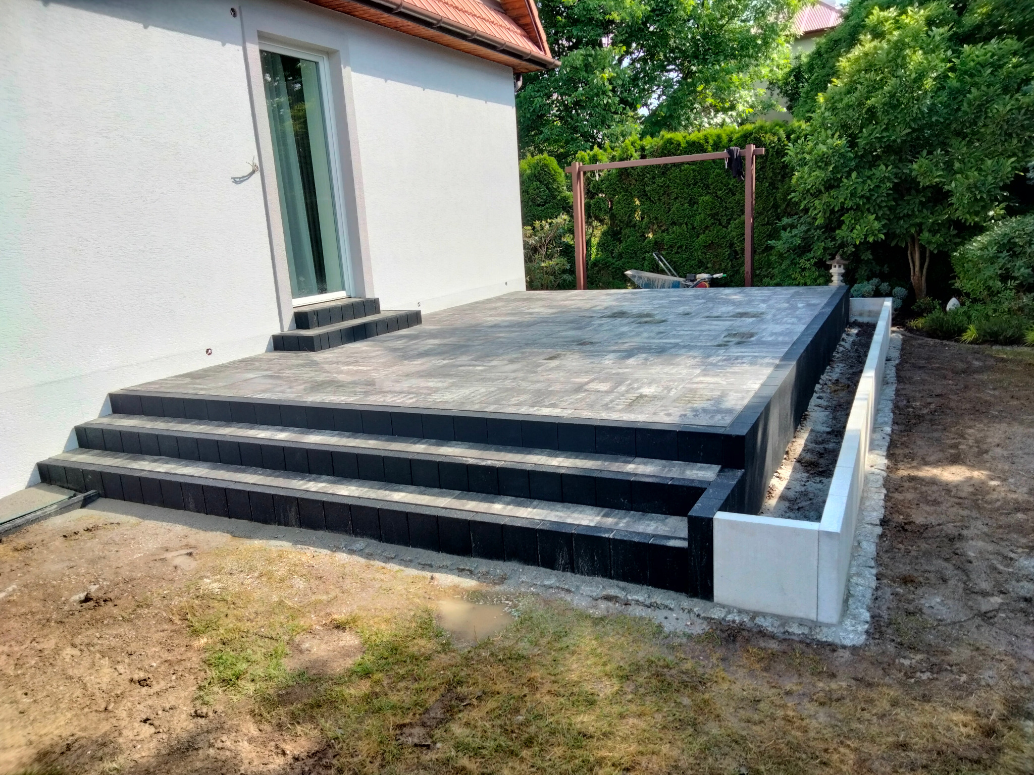 Nowoczesny taras z kostki brukowej w odcieniach szarości i czerni, z trzema stopniami i betonową obudową rabaty, przylegający do białego budynku z oknem.