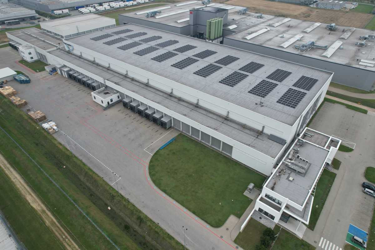 Widok z lotu ptaka na duży, biały budynek magazynowy z panelami słonecznymi na dachu, widoczne rampy załadunkowe, teren zielony i palety na zewnątrz.