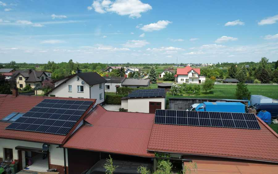 Widok z góry na kilka domów jednorodzinnych z panelami słonecznymi na dachach, w tle zielone pola i błękitne niebo z chmurami.