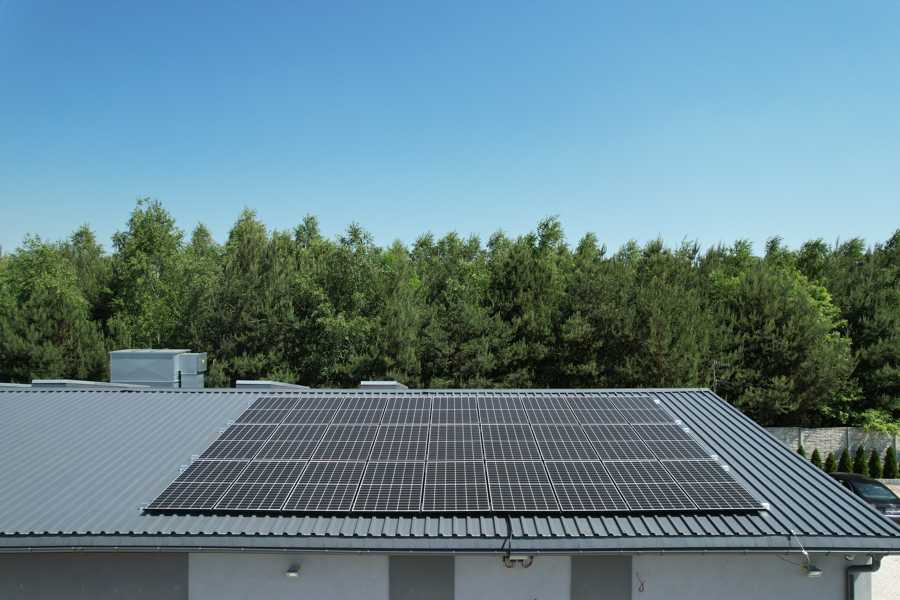 Panel słoneczny na dachu budynku z blachy trapezowej, widok z góry, tło stanowi las i błękitne niebo.