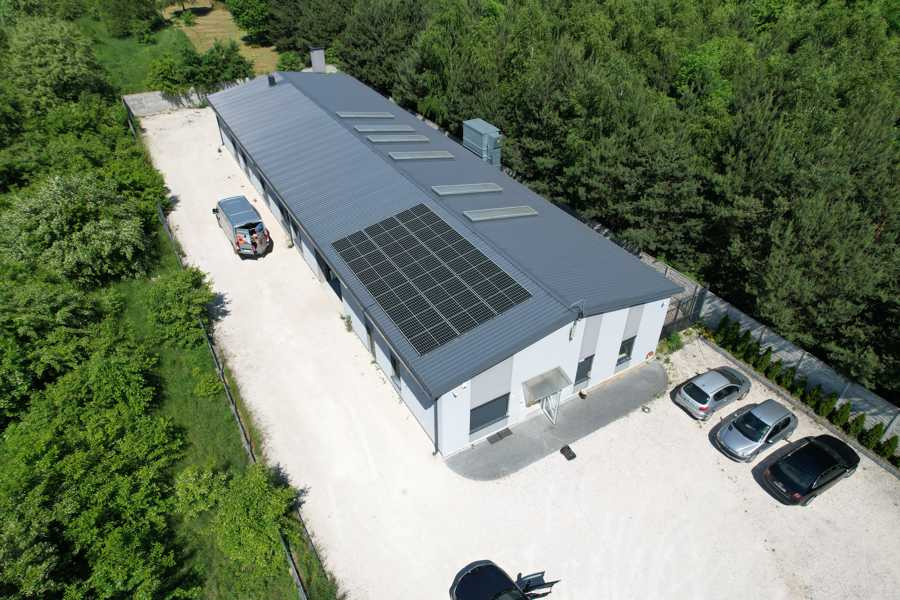 Widok z lotu ptaka na budynek z panelami słonecznymi na dachu, otoczony drzewami i zaparkowanymi samochodami na żwirowym parkingu.