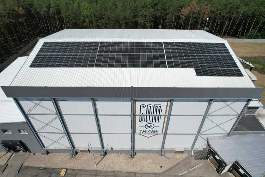 Widok z góry na duży budynek z panelami słonecznymi na dachu i logo firmy na ścianie, otoczony drzewami.