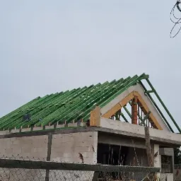 Konstrukcja dachu z zielonymi łatami na surowym budynku, widok z dołu na tle pochmurnego nieba i słupa energetycznego.